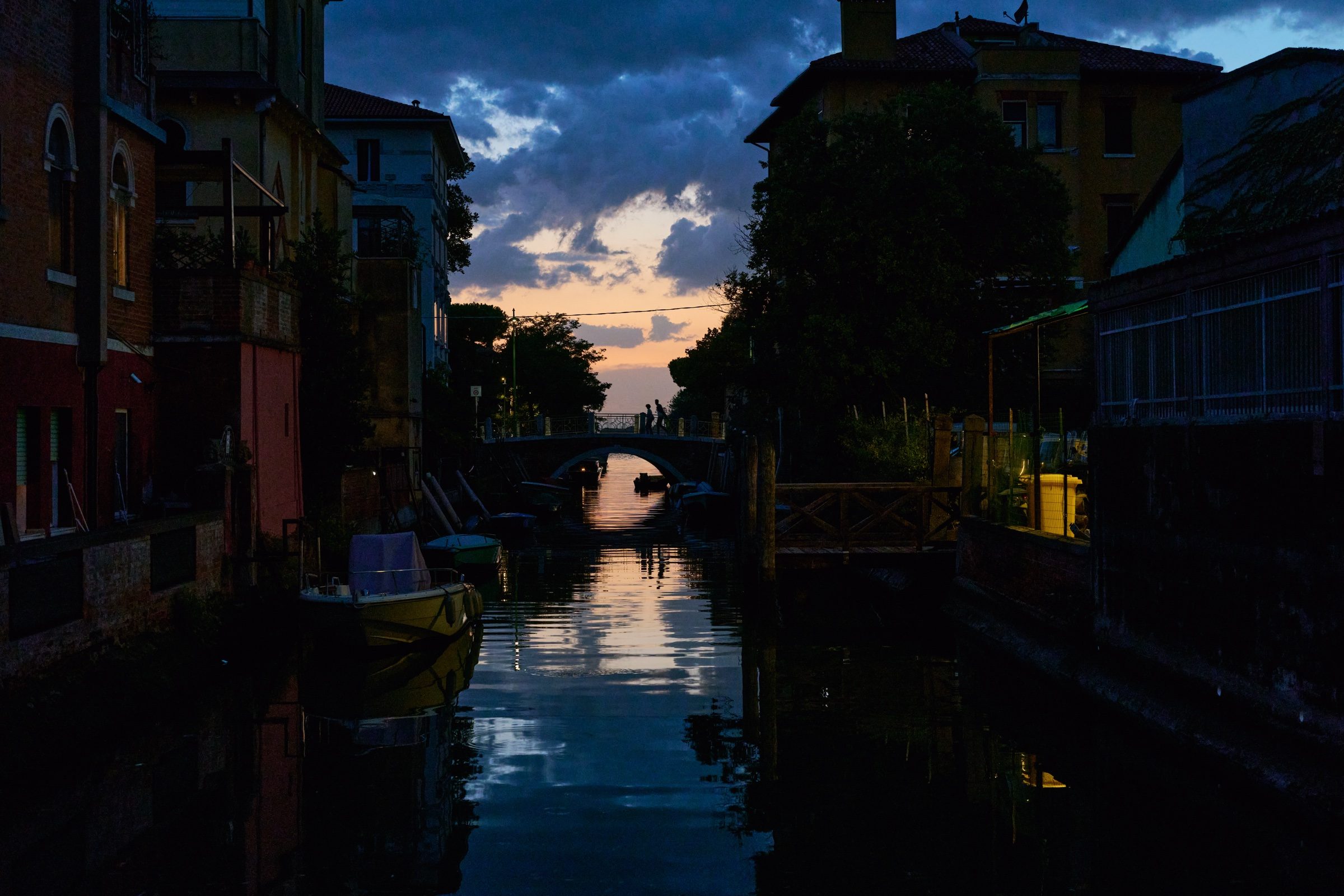 A narrow canal at dusk, houses and a bridge reflected in the still water under a glowing sky, Venice