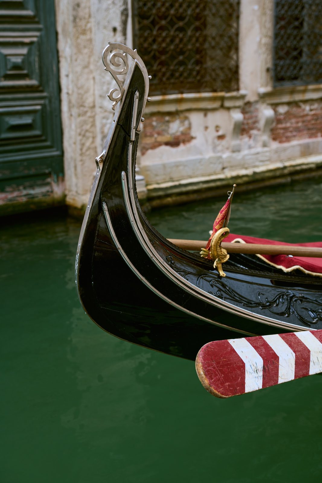 The curled iron prow and red-and-white mooring post of a gondola on green water, Venice