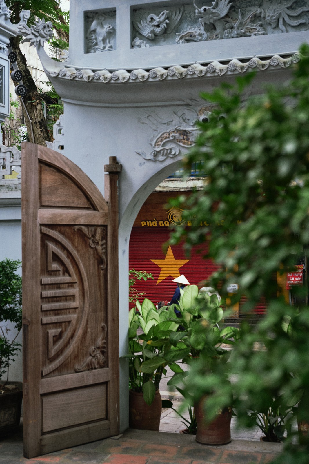 Red shutter painted with a yellow star seen through a white archway as a person in a conical hat passes, Hanoi, Vietnam