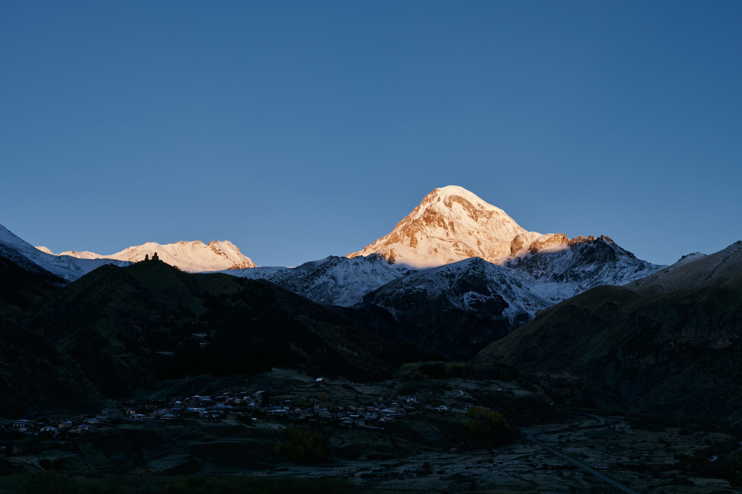 Mount Kazbek glowing in early sun above the shadowed valley, with Gergeti Trinity Church on the ridge, Georgia