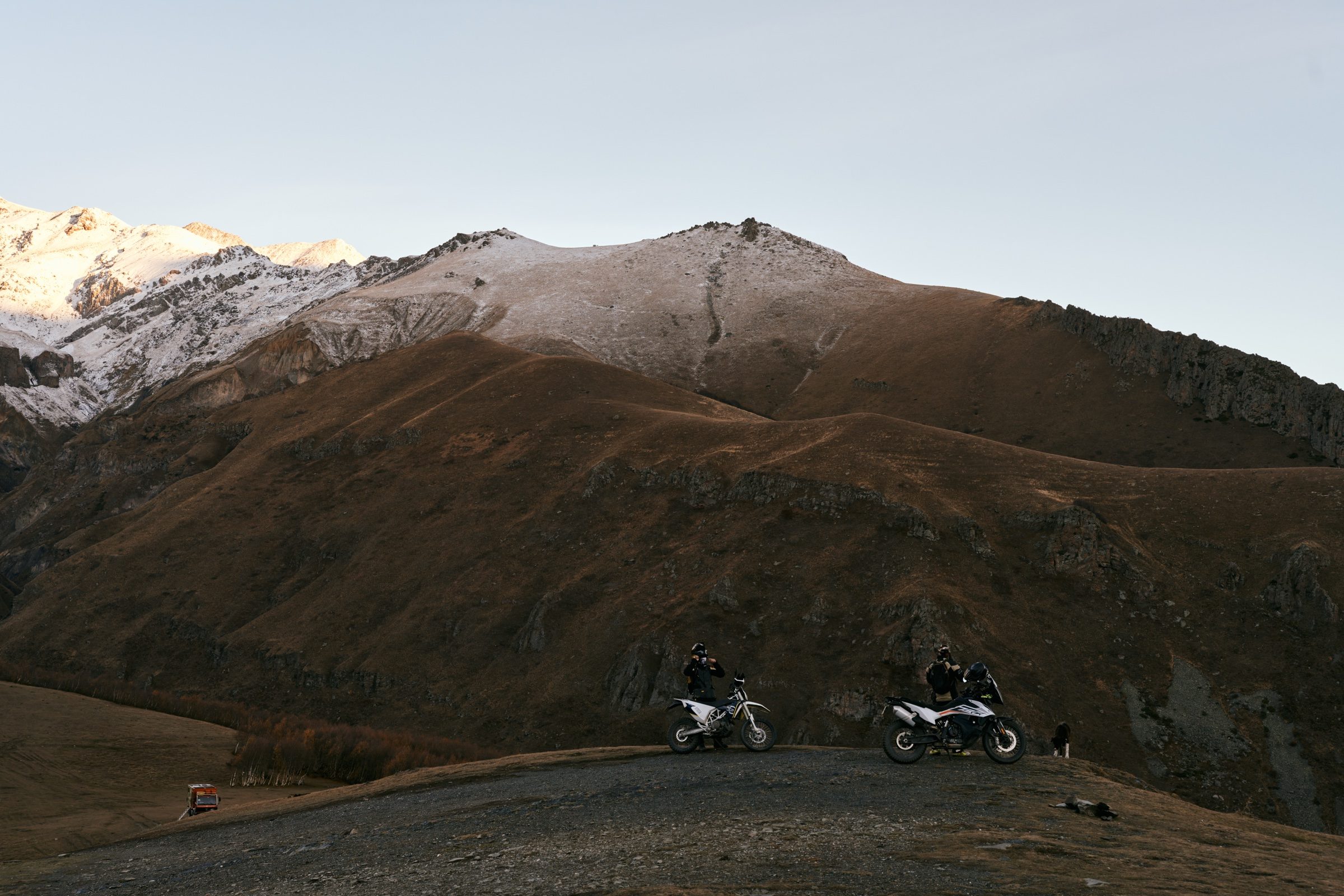 Two motorcycles parked on a high gravel pass beneath snow-dusted brown mountains near Kazbegi, Georgia