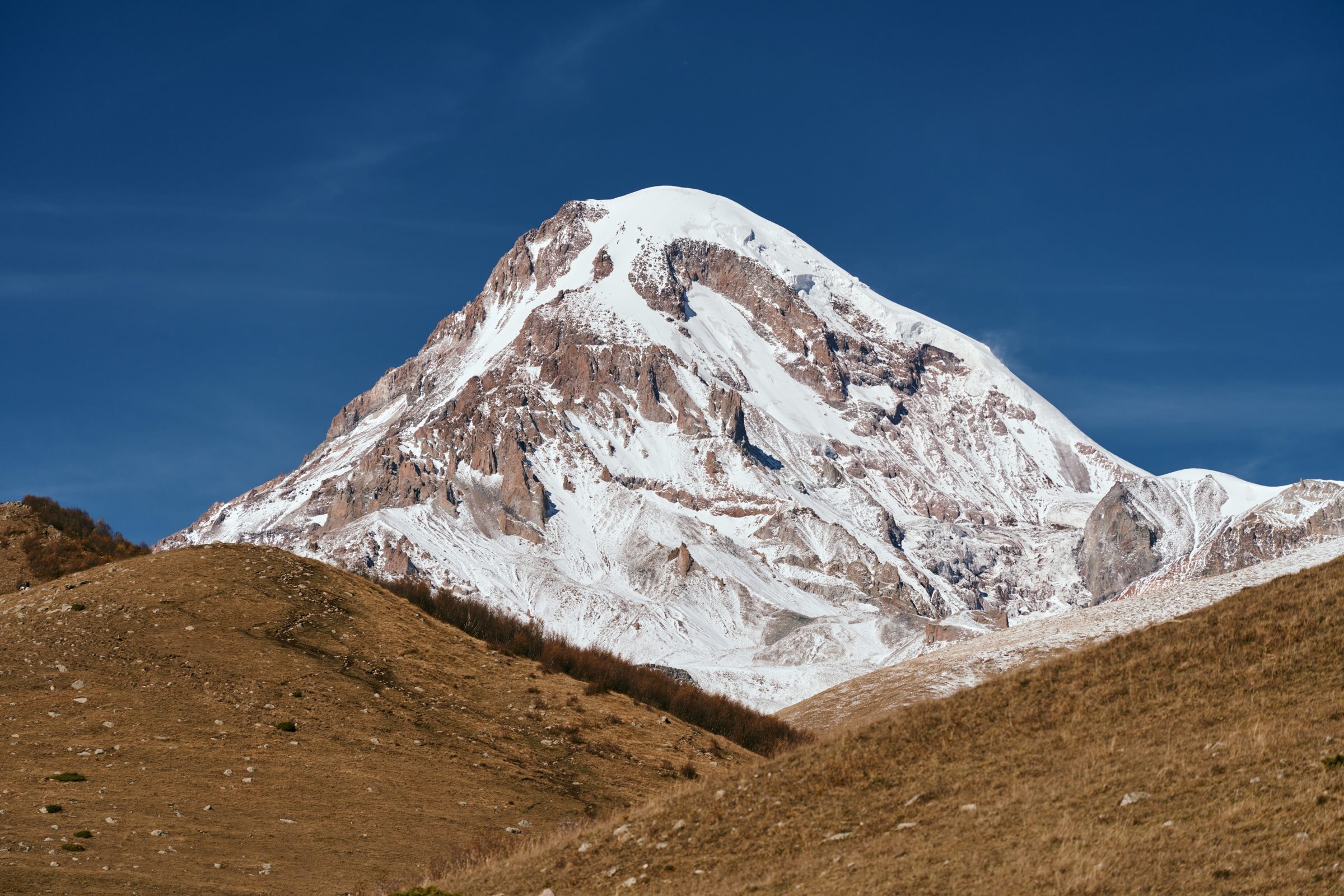 Close view of Mount Kazbek's snowy, rust-streaked summit above brown hills, Georgia