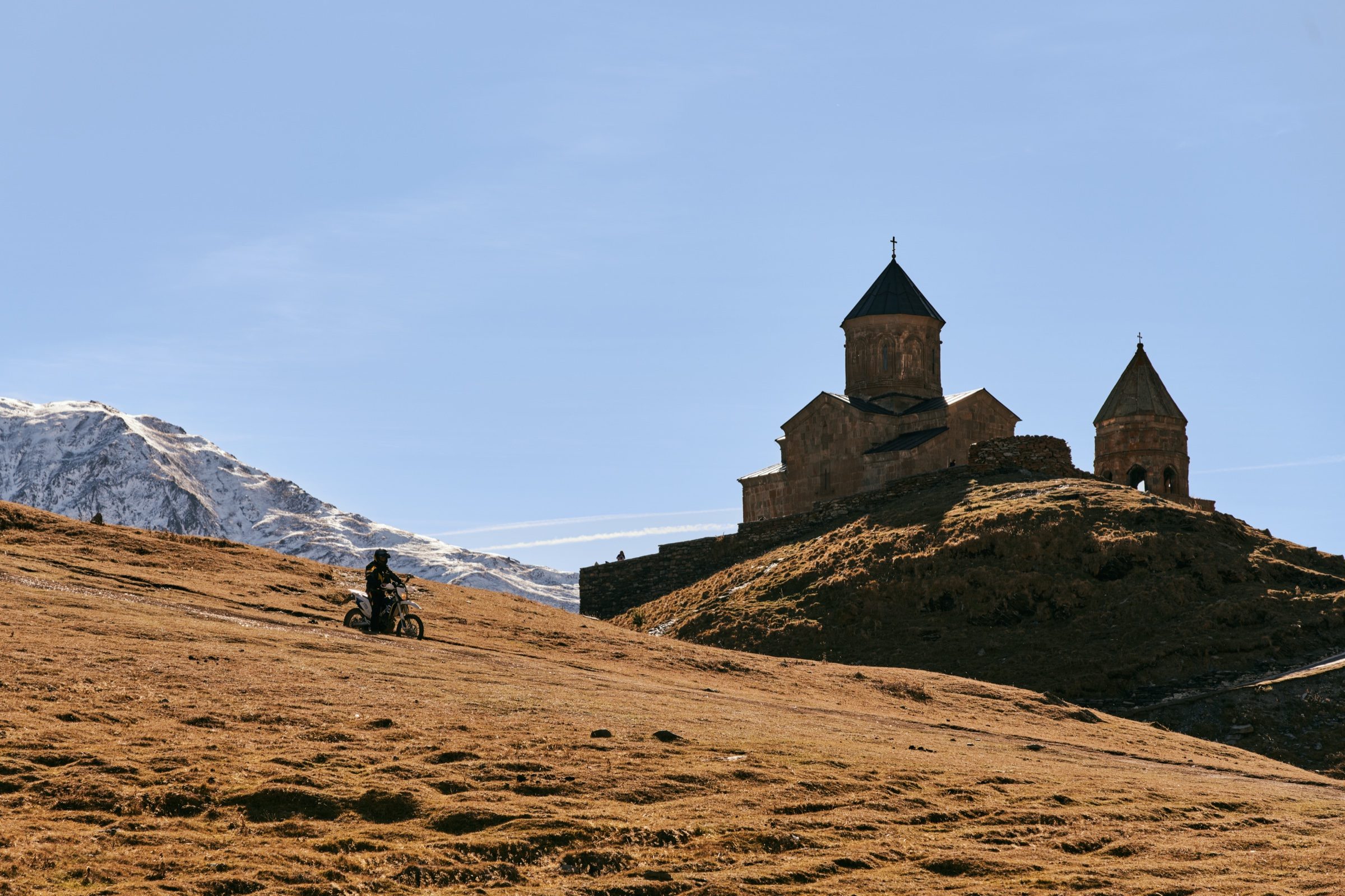A lone rider on a golden hillside below Gergeti Trinity Church, snowy peaks behind, Georgia