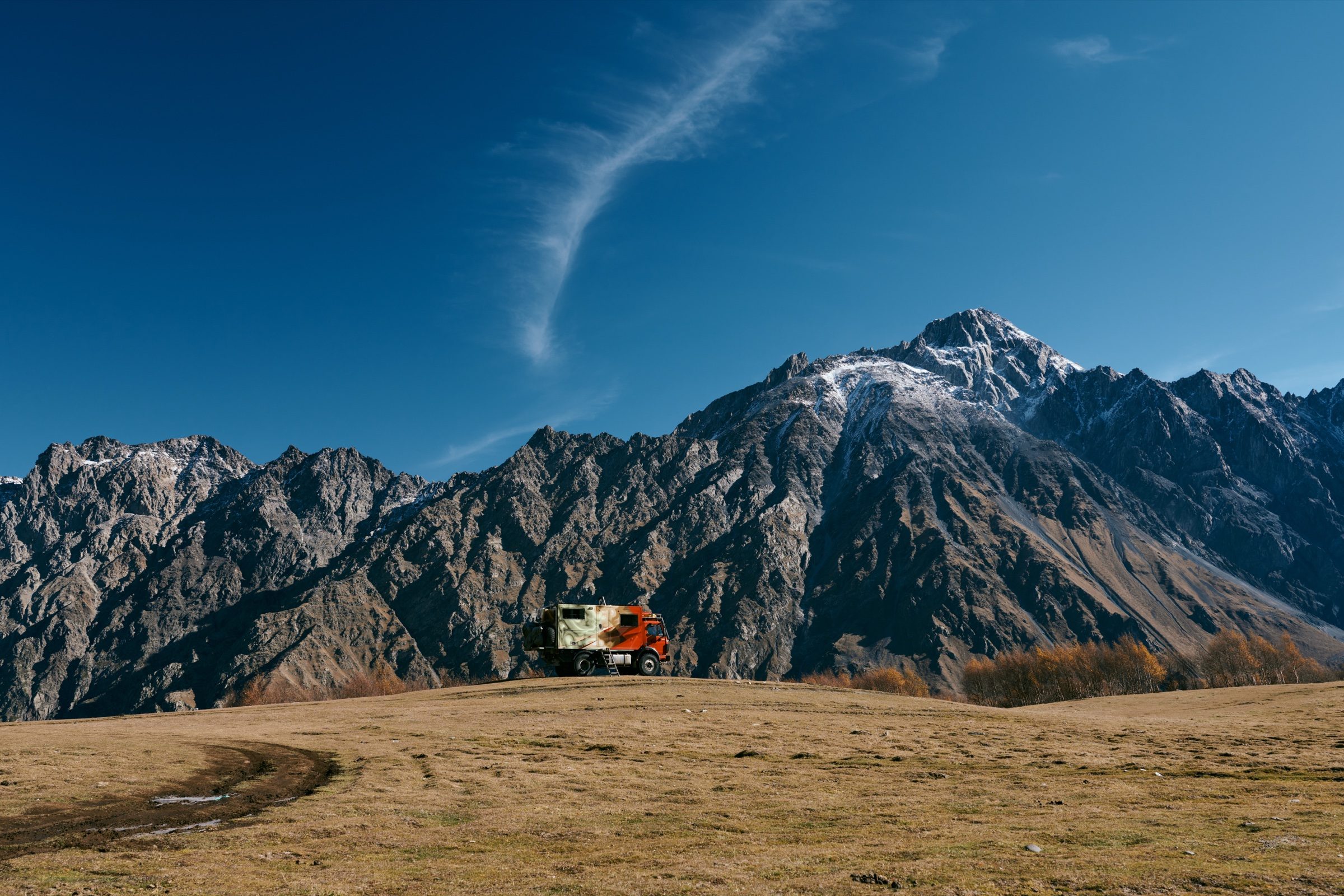 A camper van on a dry grassy plateau below jagged mountains, with a contrail across the blue sky, Georgia
