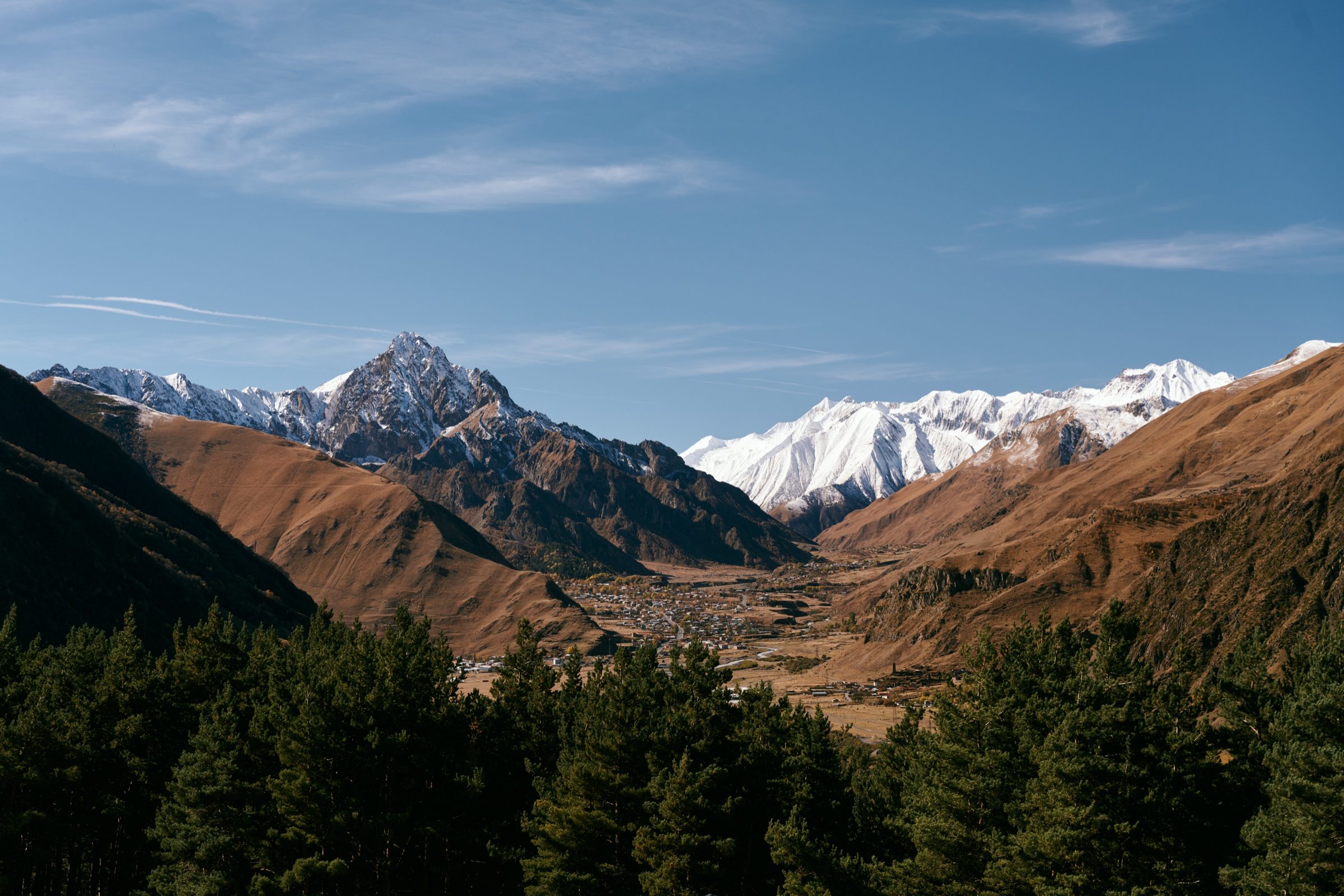 View down a forested valley toward snowy peaks under wispy clouds, Kazbegi, Georgia