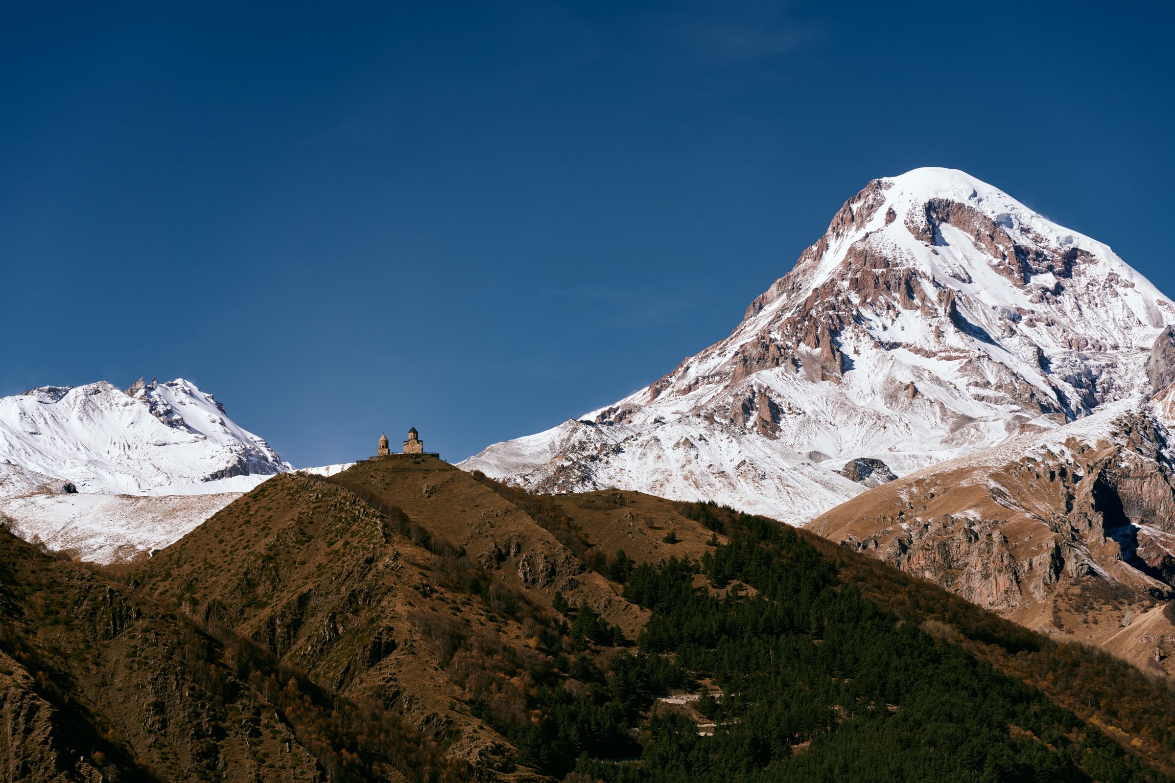 Gergeti Trinity Church tiny on a forested ridge below the towering snowy peak of Mount Kazbek, Georgia