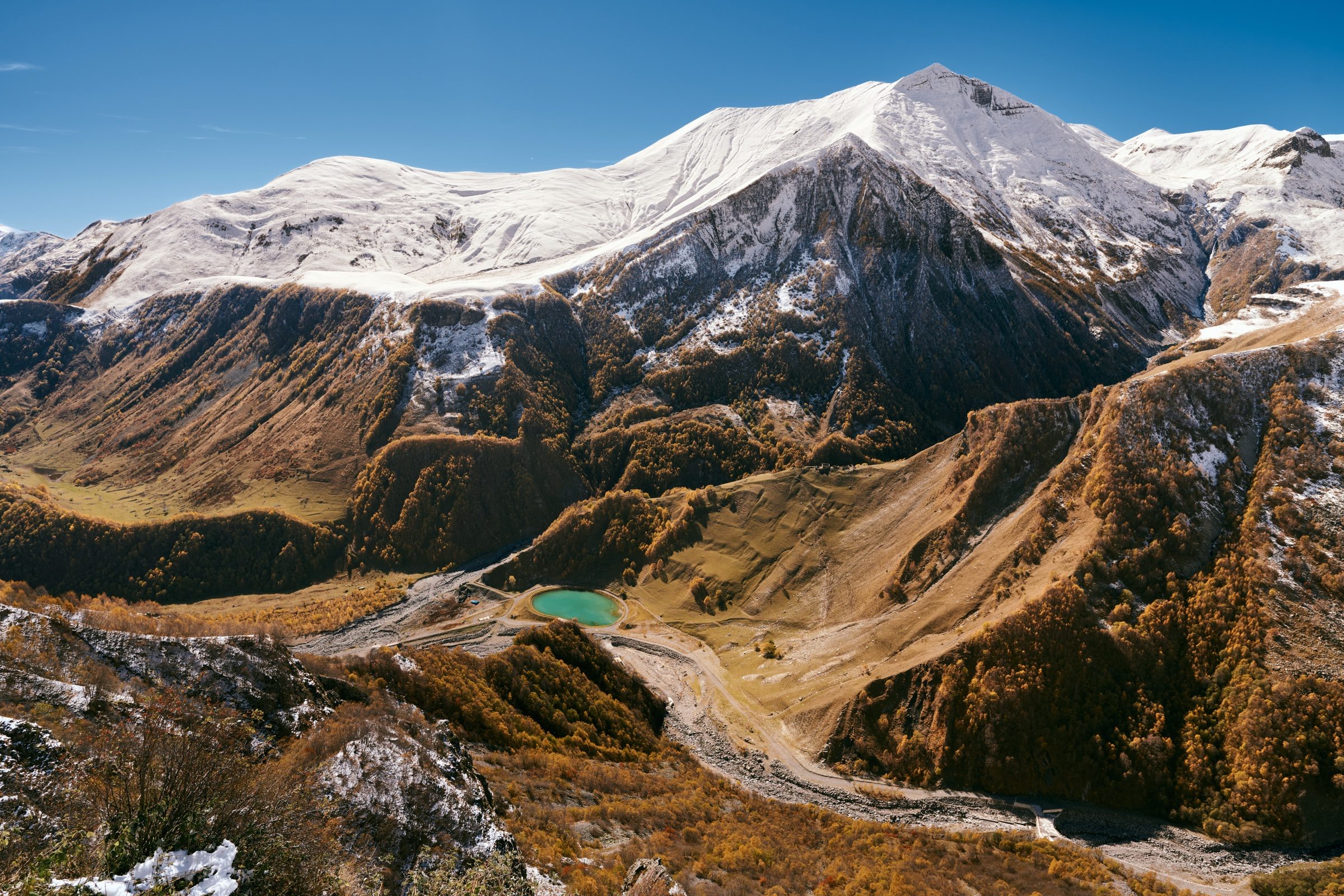 Winding mountain road and a small turquoise lake below snow-capped peaks in autumn colours, Georgia