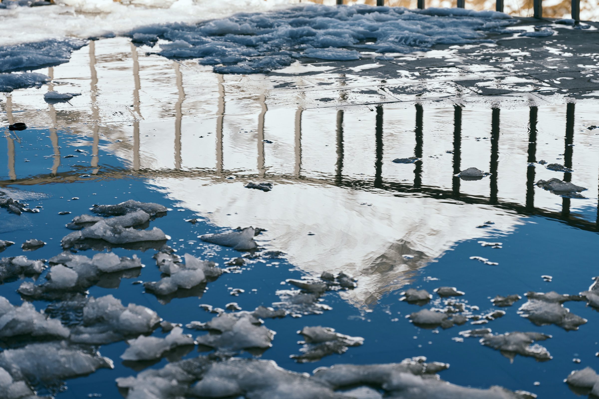 Railings and snowy mountains reflected in icy meltwater at the Friendship Monument, Georgia