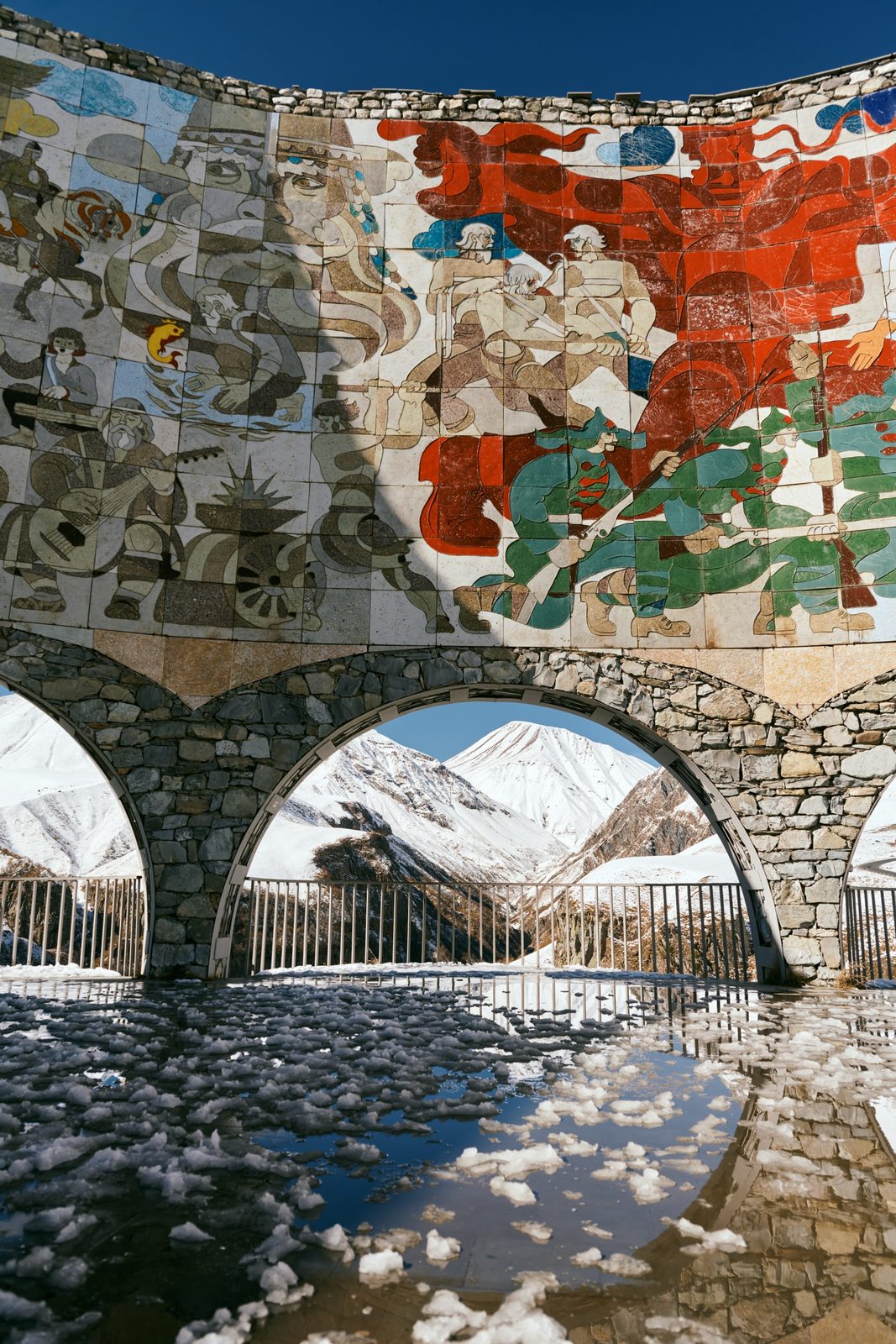 Mosaic wall above stone arches framing snowy peaks, mirrored in melting ice, Friendship Monument, Georgia