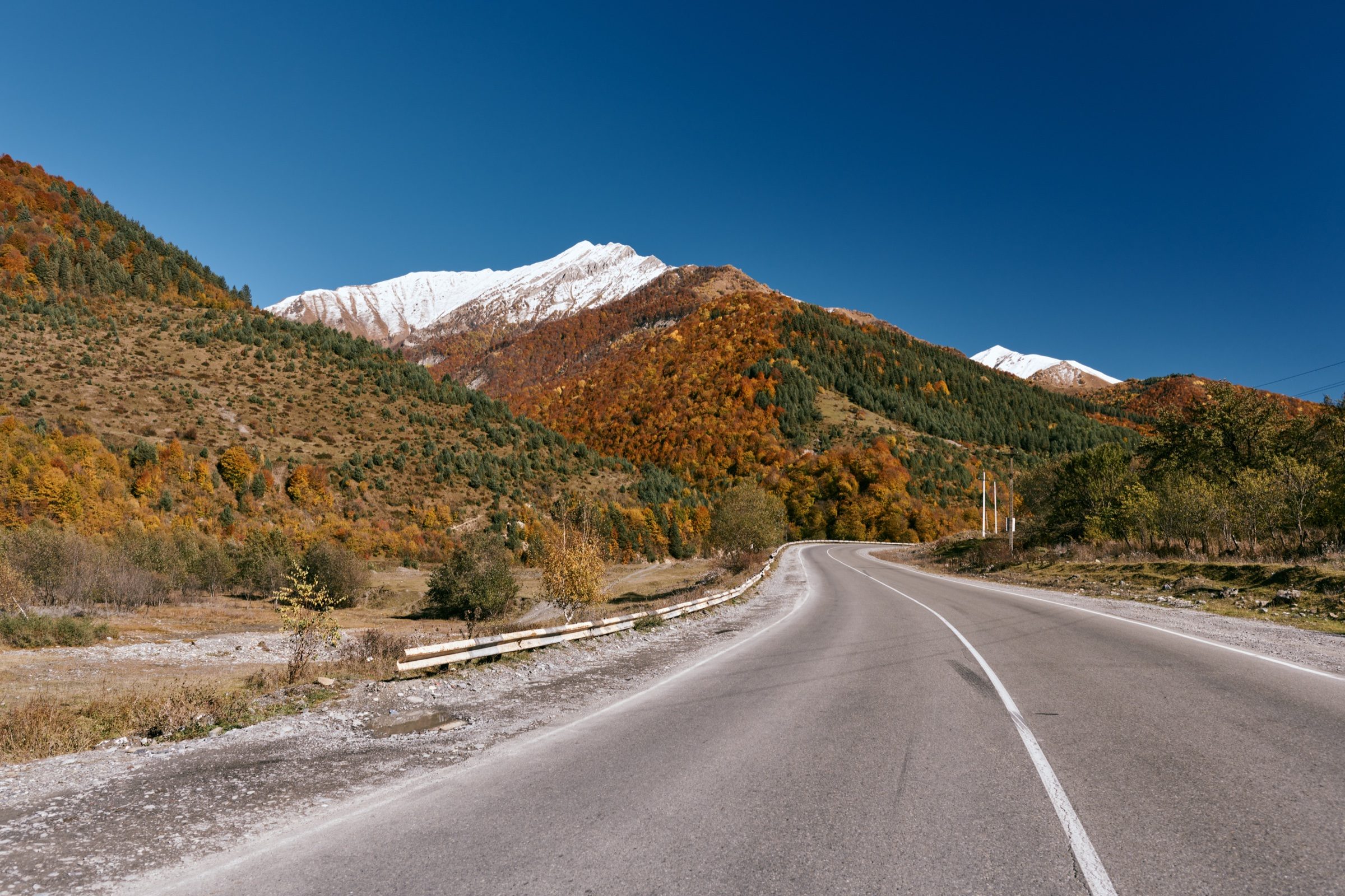 An empty road curving through autumn hills toward snow-capped mountains on the way out of Kazbegi, Georgia
