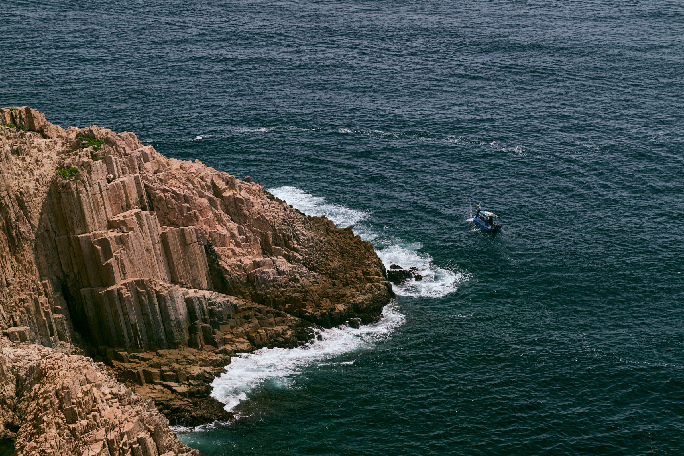 Hexagonal volcanic rock columns meeting surf, with a boat offshore, MacLehose Trail, Sai Kung, Hong Kong