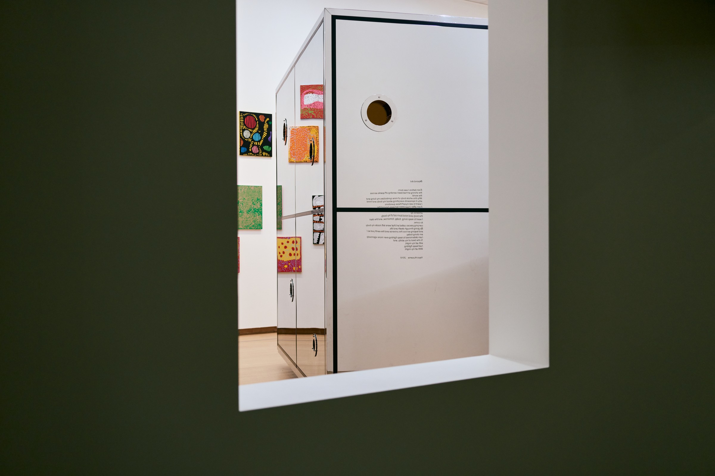 View through a wall opening to a white installation box with a round peephole, printed with Kusama's poem 'Beyond Art'