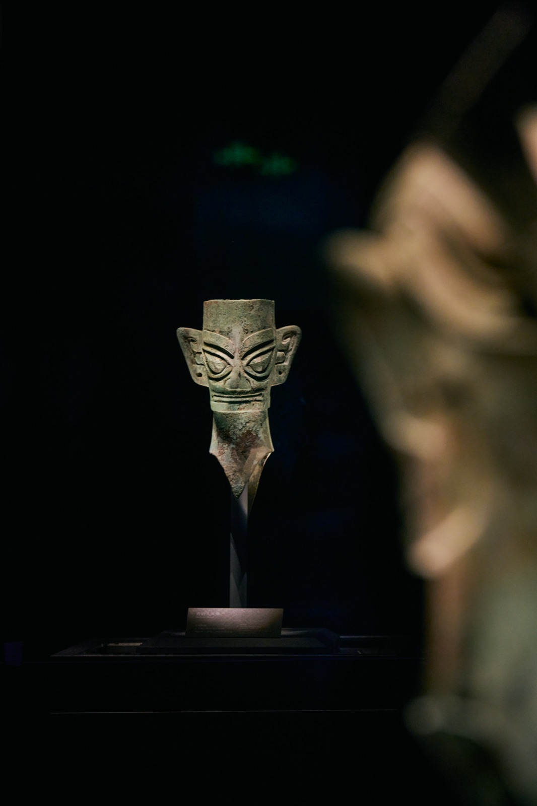 Small bronze head lit in the distance, a large head blurred in the foreground, Sanxingdui Museum, Guanghan, China
