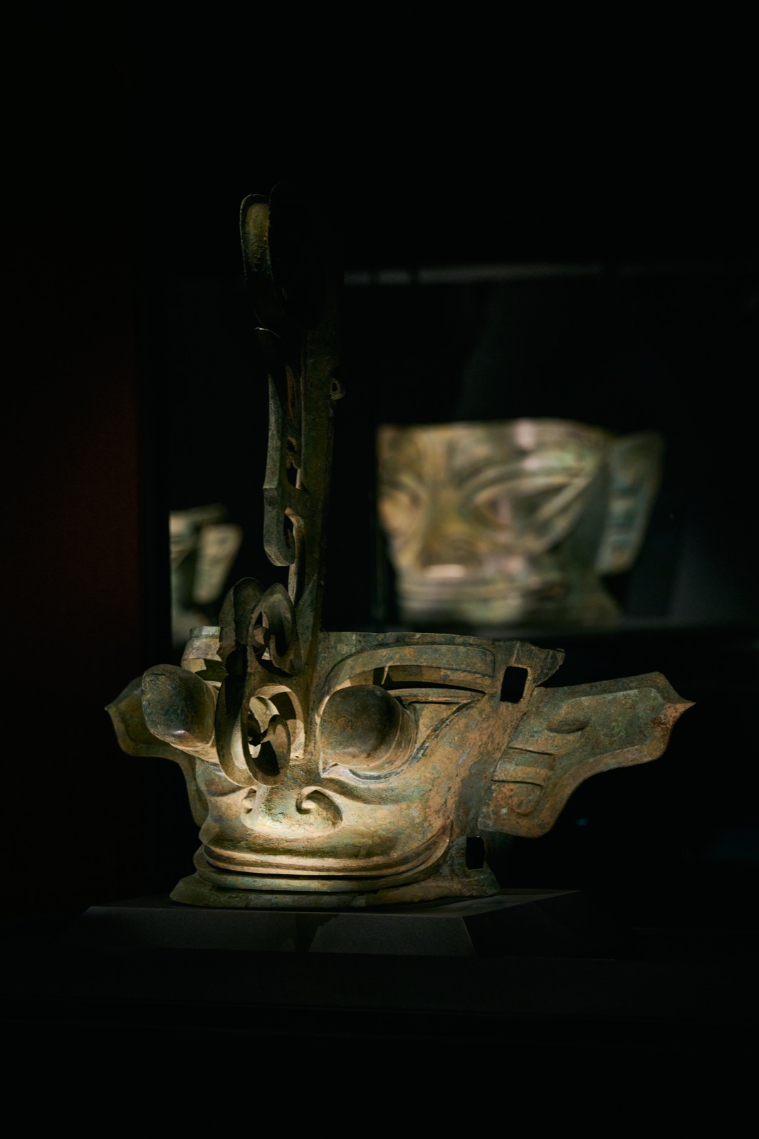 Bronze mask with a tall curled forehead ornament, another mask blurred behind, Sanxingdui Museum, Guanghan, China